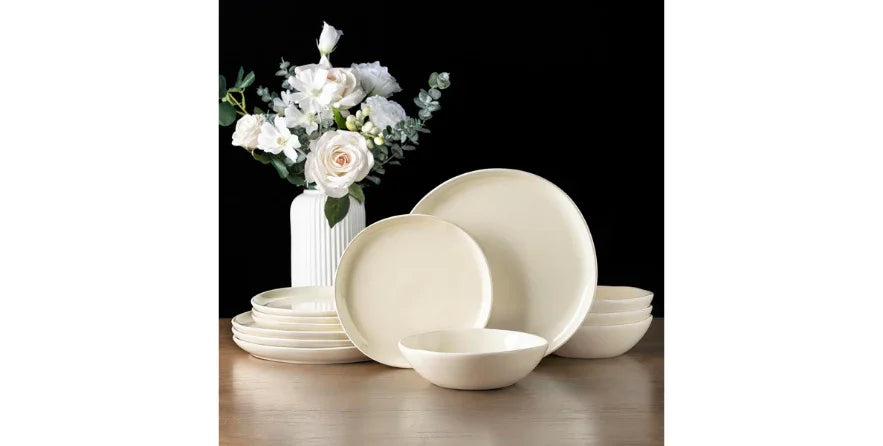 Cream-colored ceramic dinnerware set with plates and bowls on wooden table, white flower vase backdrop