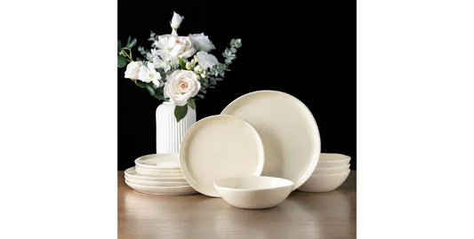 Cream-colored ceramic dinnerware set with plates and bowls on wooden table, white flower vase backdrop