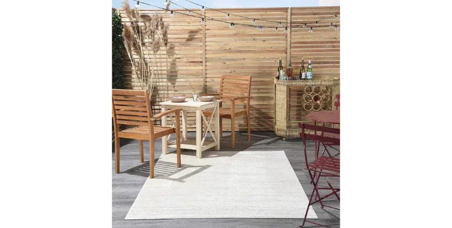 Ivory beige indoor outdoor rug on patio with wooden chairs, table, and wicker bar against wooden fence