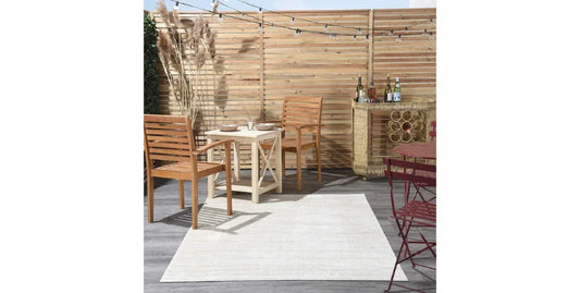 Ivory beige indoor outdoor rug on patio with wooden chairs, table, and wicker bar against wooden fence